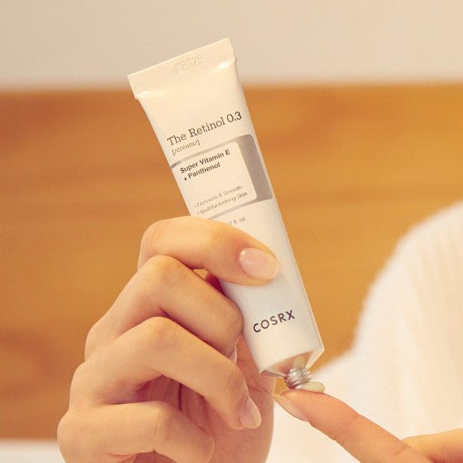 Hand squeezing pale cream from tube labeled Retinol 0.3 with vitamin E and panthenol for wrinkle reduction benefits