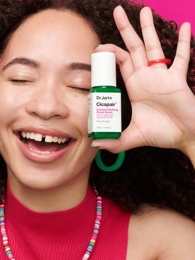 Woman smiling and holding Dr.Jart+ Cicapair Intensive Soothing Repair Serum 30ml bottle near her face against pink background