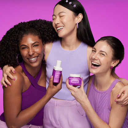 Three smiling women in purple outfits holding Dr.Jart+ Prejuvenation Firming Bakuchiol Cream 50ml against pink background