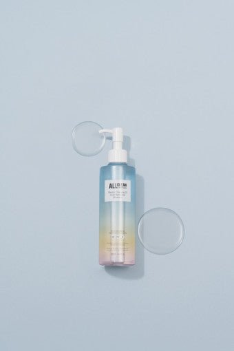 Clear gradient bottle of cleansing oil with pump dispenser on pale blue surface alongside two clear liquid drops