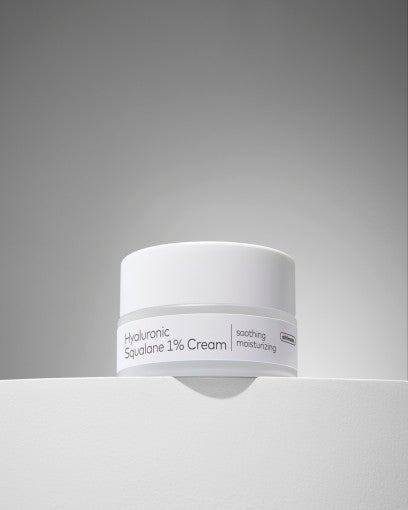 White jar of soothing moisturizing cream labeled Hyaluronic Squalane 1% on smooth pale surface with gray background