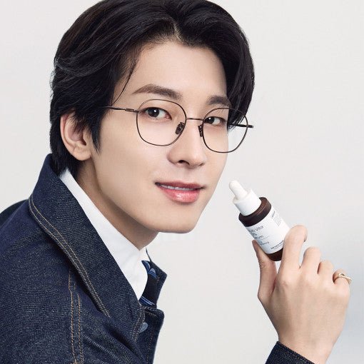 Man with glasses smiling while holding a small brown dropper bottle serum against a plain light background