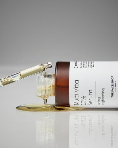 Glass dropper releasing light yellow serum onto reflective surface beside amber serum bottle labeled Multi Vita 10% brightening