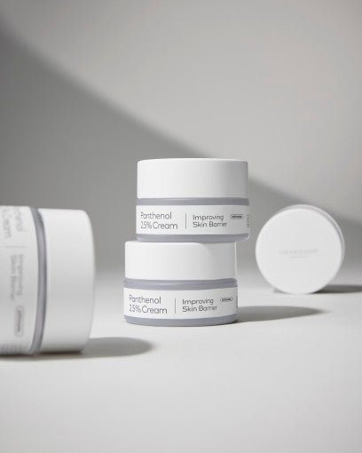 Two white jars of panthenol cream stacked on light surface with soft shadows and a blurred jar lid nearby