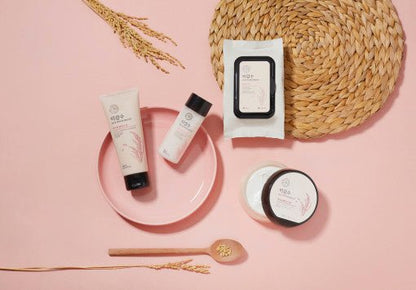 Collection of skincare products with rice-inspired packaging arranged on pink surface with woven placemat and spoon