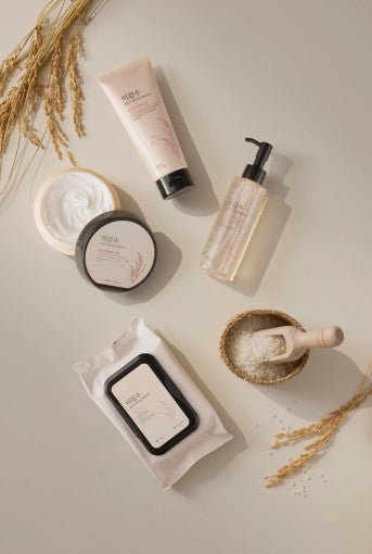Set of skincare products including cleansing wipes, cream jars, and pump bottle with dried rice stalks and bowl of rice grains