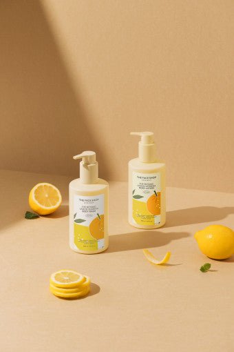 Two cream-colored lotion bottles with lemon label and pump tops surrounded by fresh lemon slices and leaves on beige surface