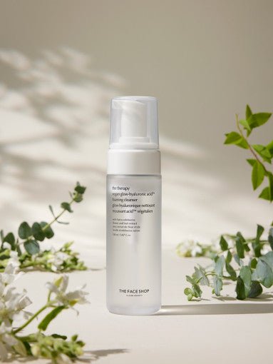 Tall clear pump bottle of foaming cleanser with text label placed among green foliage on beige background