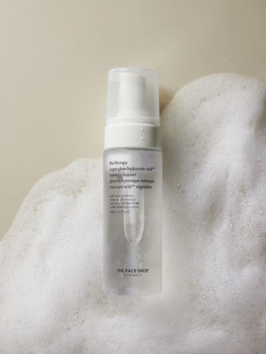Clear pump bottle of hyaluronic acid foaming cleanser surrounded by light, airy white foam on beige background