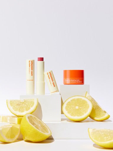 Lip balm stick with vibrant color beside bright orange moisturizer jar and fresh lemon slices on white blocks