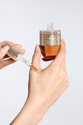 Hand holding amber glass ampoule bottle with dropper dispensing clear serum onto wrist against white background