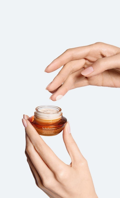 Hands with neatly manicured nails holding and applying a small amber jar of hydrating eye cream against a white background