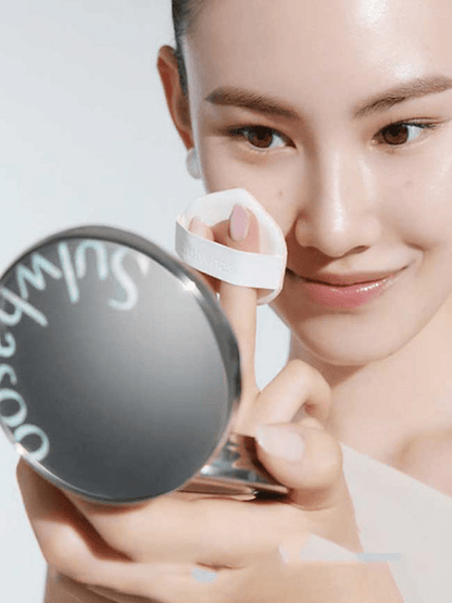 Woman with clear skin using a white cushion puff to apply lightweight cushion foundation while holding a compact mirror