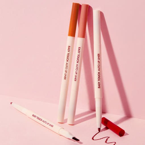 Three slim automatic lip liners in warm shades standing on pink surface with one uncapped showing fine tip and drawn line