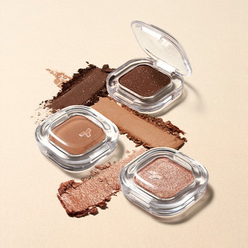 Three single eyeshadows in clear square cases with soft-textured shades in brown, beige, and shimmery rose tones on beige background