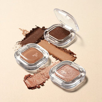 Three single eyeshadows in clear square cases with soft-textured shades in brown, beige, and shimmery rose tones on beige background