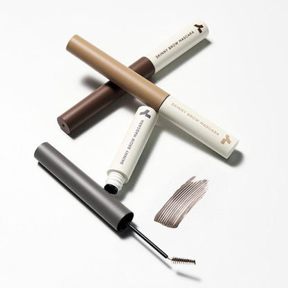 Three slim mascara tubes in neutral tones with a small brow brush and a swatch of light brown product nearby