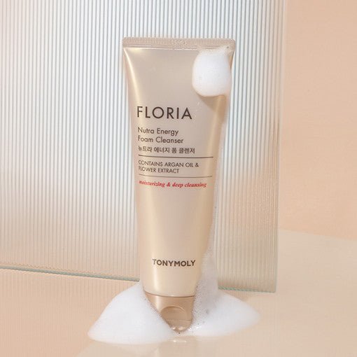 Beige tube of foam cleanser with argan oil and flower extract text, surrounded by white foam bubbles on soft surface
