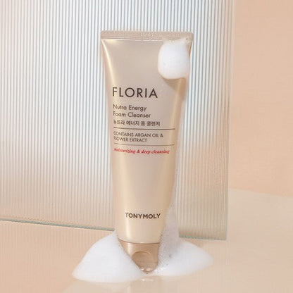Beige tube of foam cleanser with argan oil and flower extract text, surrounded by white foam bubbles on soft surface