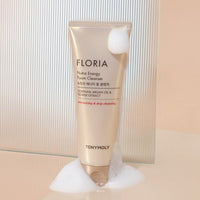 Beige tube of foam cleanser with argan oil and flower extract text surrounded by white foam on soft background