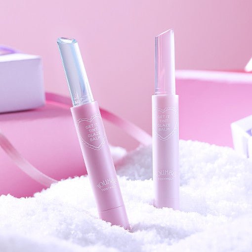 Two pastel pink lip balms with clear caps standing upright on white fluffy surface against soft pink background