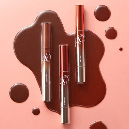 Three sleek lip balms with glossy brown tint spilled artistically on soft peach background