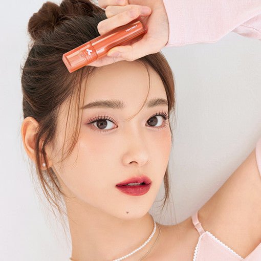 Young woman with natural makeup holding coral-colored long-lasting lip tint near forehead against white background