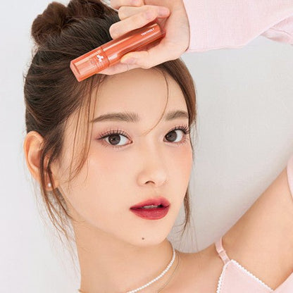 Young woman with natural makeup holding coral-colored long-lasting lip tint near forehead against white background