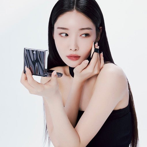 Woman with long dark hair applying cushion foundation using a compact mirror against a plain background