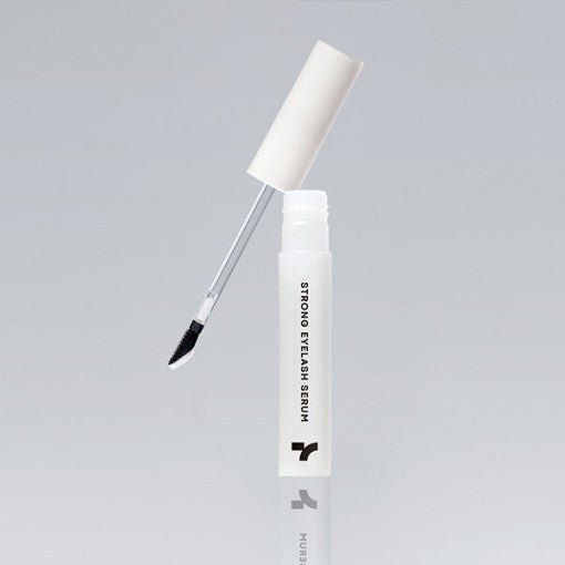 Open white bottle of eyelash serum with angled applicator brush against plain gray background