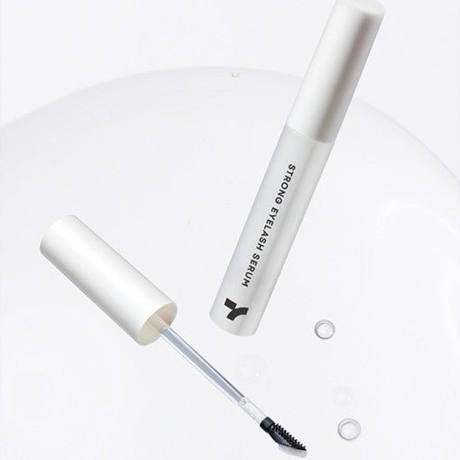 Minimalist white tube of eyelash serum with applicator brush and clear serum droplets on a smooth white surface
