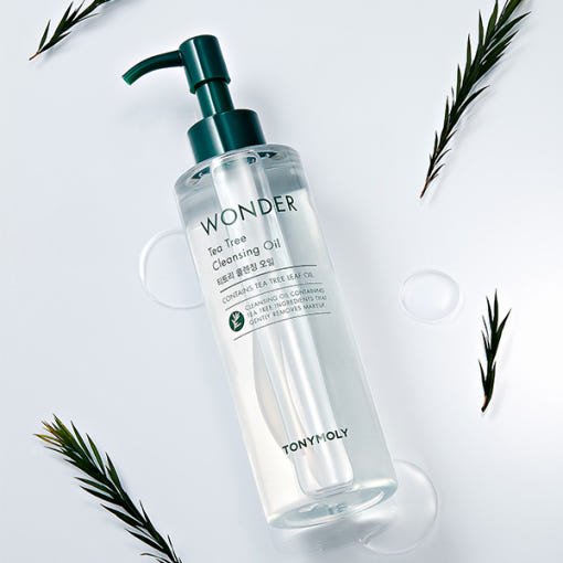 Clear pump bottle of tea tree cleansing oil with dark green cap surrounded by fresh tea tree leaves on white surface