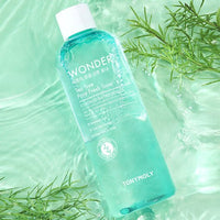 Clear blue toner bottle with tea tree extract placed on water surface surrounded by green leaves