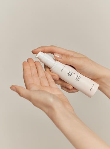 Hand holding white pump bottle of Ginseng Moist Sun Serum dispensing product onto palm on beige background