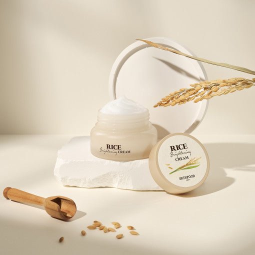 Open jar of hydrating rice brightening cream with beige lid and wooden scoop on pale background with rice grains