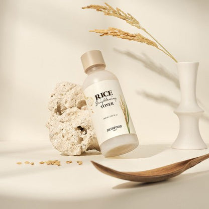 Bottle of nourishing rice brightening toner resting against textured stone with dried rice stalks in white vase nearby