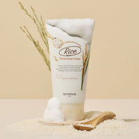 Tube of brightening rice cleansing foam with foamy lather, surrounded by rice grains and wooden spoon on beige surface