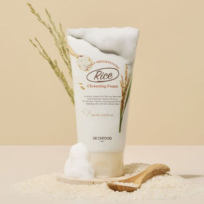 Tube of brightening rice cleansing foam with foamy lather, surrounded by rice grains and wooden spoon on beige surface