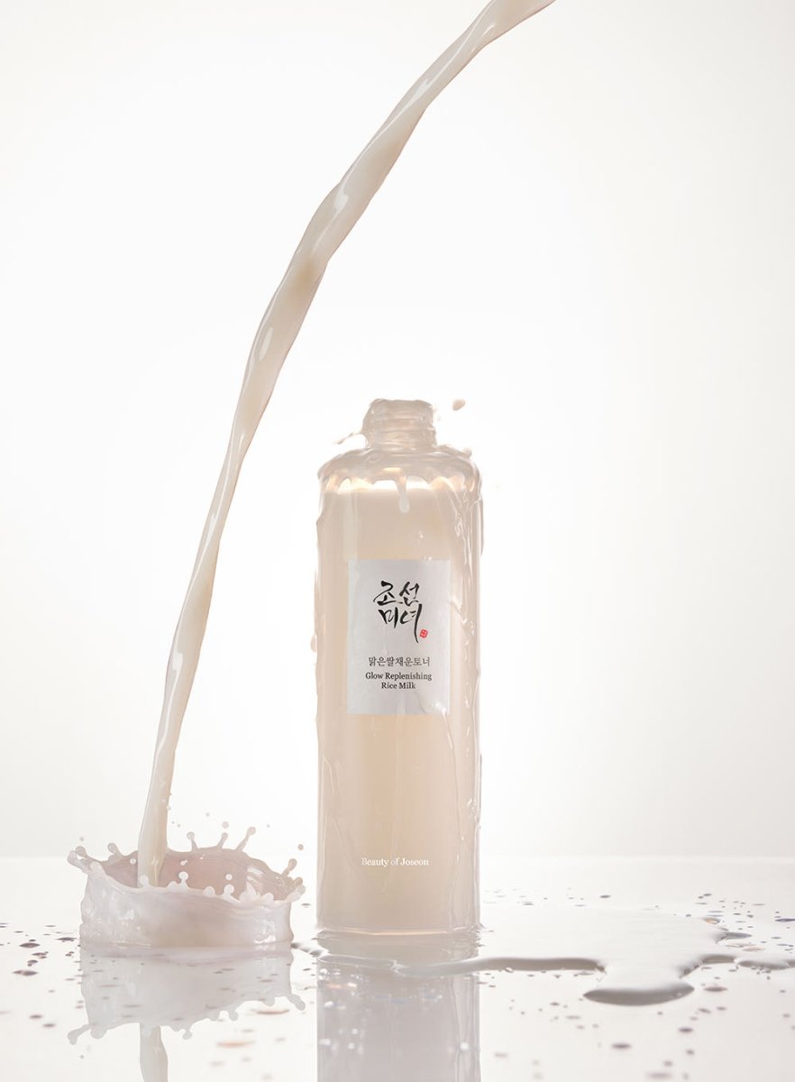 Creamy liquid flowing into an open translucent bottle of Glow Replenishing Rice Milk with splash on white surface