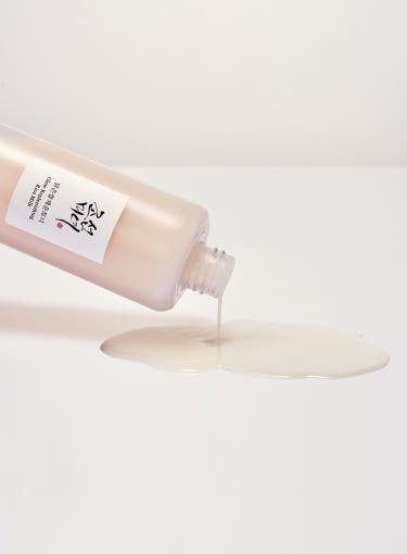 Beige translucent skincare liquid pouring from a tilted bottle onto a white surface for deep hydration effect