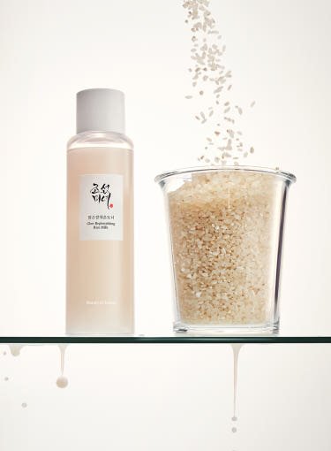 Clear bottle of Glow Replenishing Rice Milk next to a glass filled with uncooked rice grains on a white background