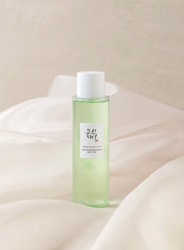 Tall translucent light green toner bottle with white cap and minimalist label on soft beige fabric background