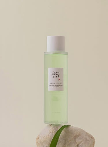 Clear green toner bottle with white cap labeled for gentle exfoliation, placed on a light stone against beige background