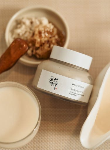 Jar of Ground Rice and Honey Glow Mask with creamy texture beside rice bowl and wooden spoon on beige surface