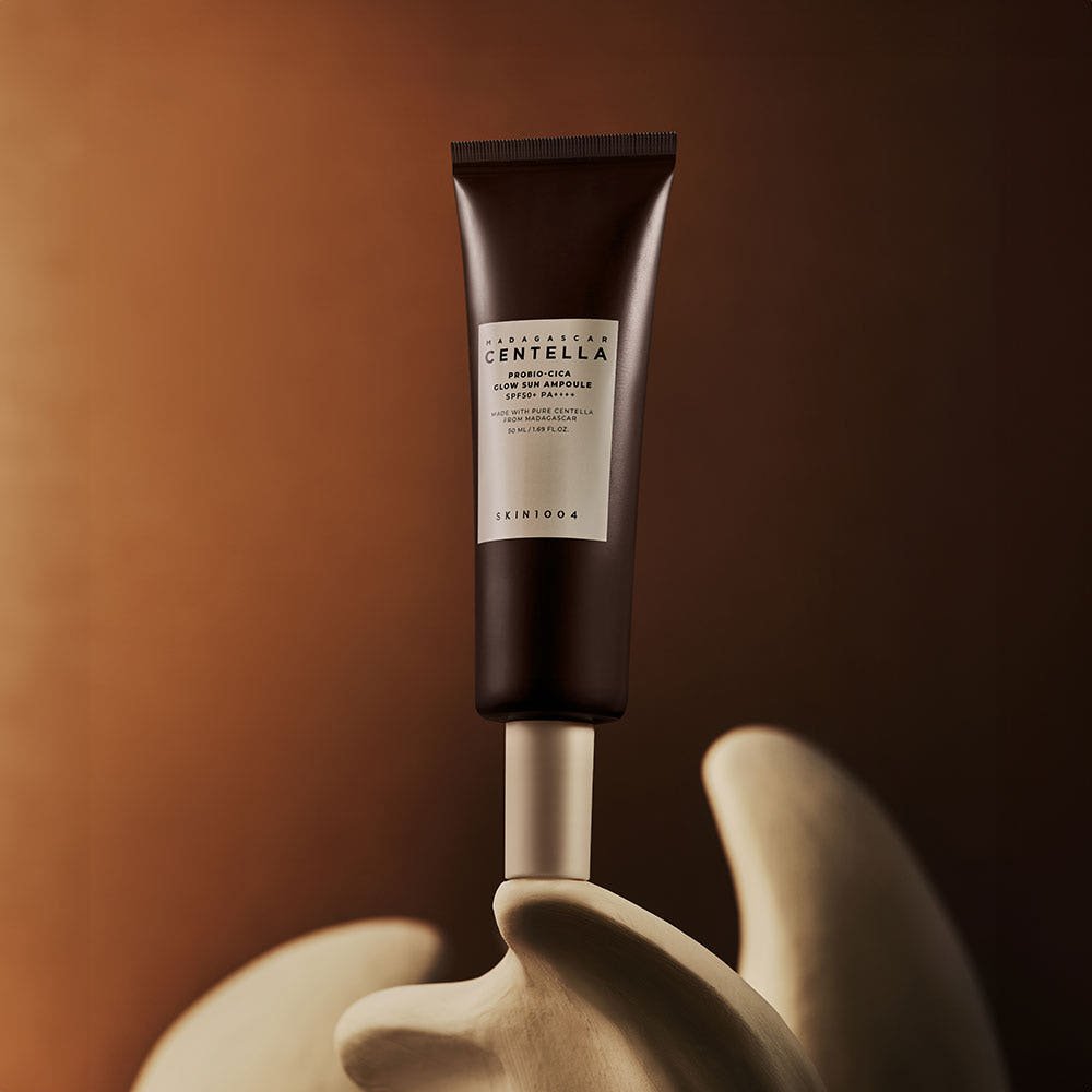 Dark brown tube of glow sun ampoule with SPF 50+ PA++++ standing on beige abstract sculpture against dark background