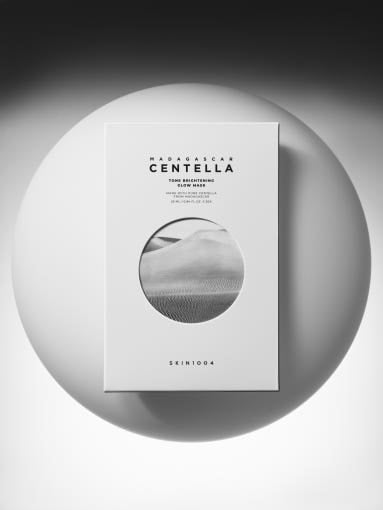 Minimalist white box packaging of brightening glow mask with a textured skin close-up on front panel against gray circular backdrop