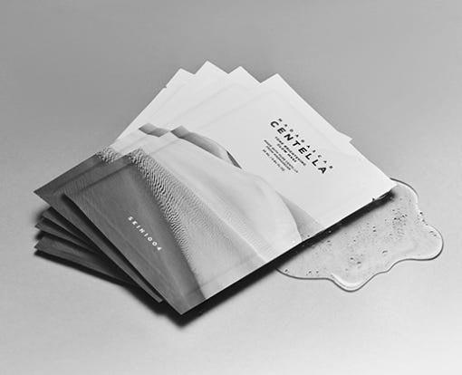 Five white sachets of brightening face masks with clear gel spilling from one packet on gray surface