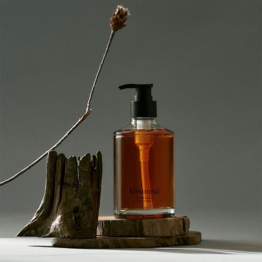 Amber liquid body wash in clear pump bottle displayed on wooden platforms with dried twig and neutral background