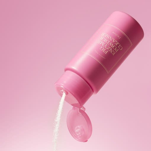Pink bottle dispensing fine white enzyme powder cleanser against a soft pink background