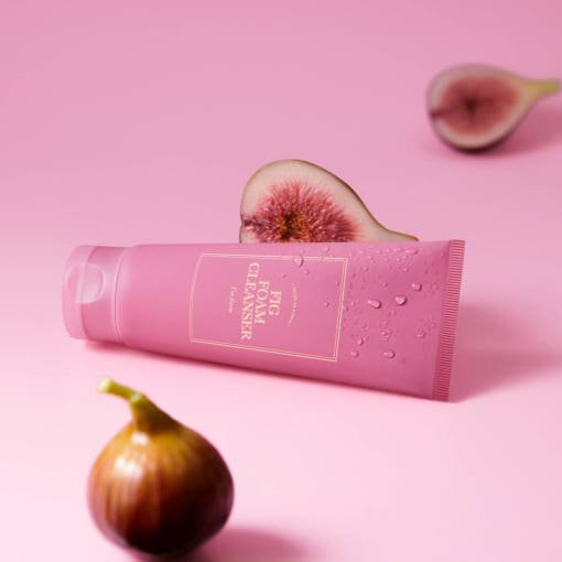 Pink tube of fig foam cleanser with water droplets beside fresh whole and halved figs on pink background